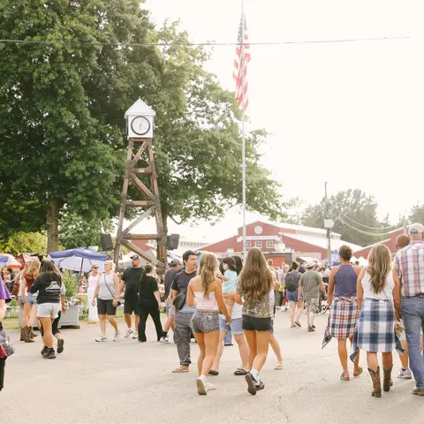 A crowd of people walking at an outdoor event near a wooden clock tower and American flag.