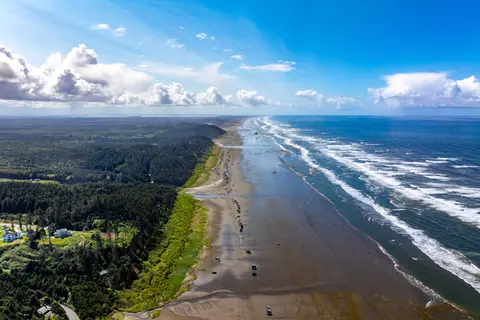 An aerial view of a wide sandy beach alongside a forested coastline and the ocean.