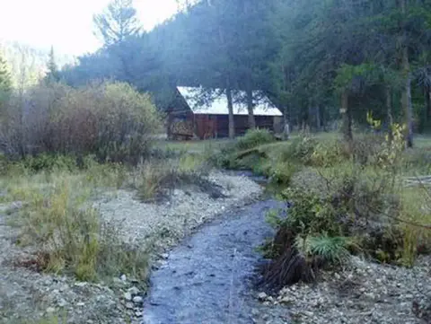 A small cabin stands near a stream in a wooded mountainous area.