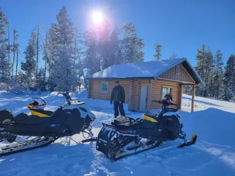 A person standing outside a log cabin in a snowy landscape with two snowmobiles.