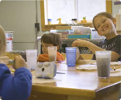 Children sit at a table painting pottery inside a room.