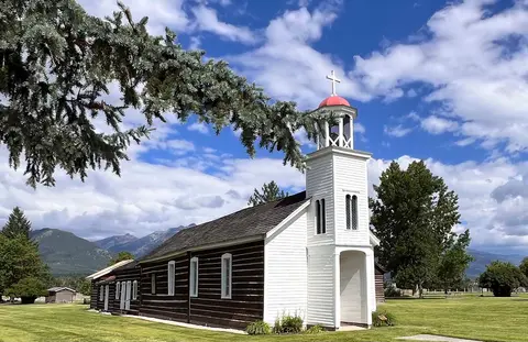An exterior view of a historic log church with a white steeple and red dome.