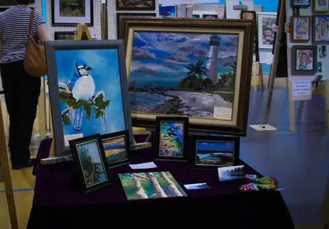 Framed paintings and art pieces are displayed on a table at an indoor art exhibit.