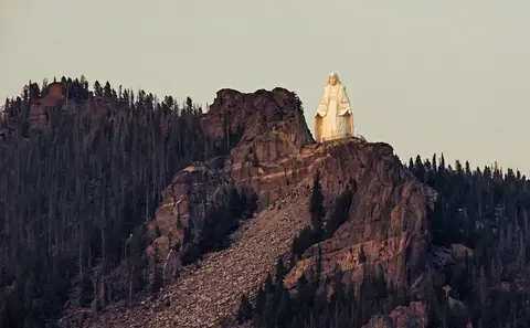 A large white statue stands on a rocky mountain ridge covered with pine trees.