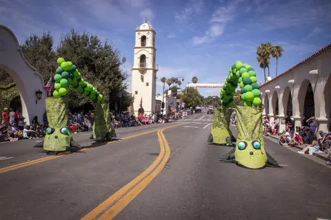 Two large green caterpillar balloon puppets are on a street during a parade with spectators watching.