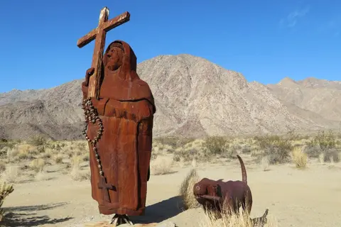 A metal sculpture of a robed figure holding a cross stands in a desert landscape with a smaller animal sculpture.