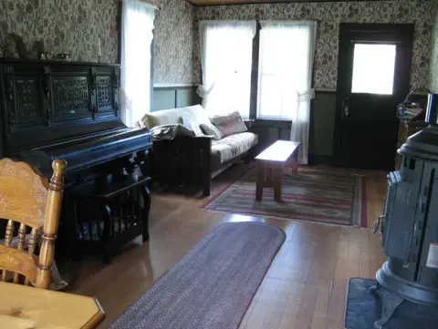 A vintage-style living room features a piano, wood stove, and patterned wallpaper.