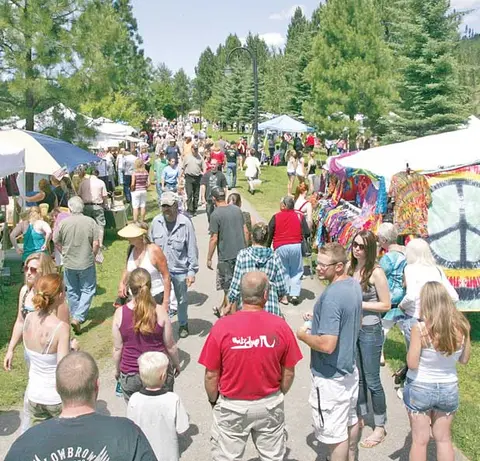 A busy outdoor street market filled with people shopping among tents and trees.
