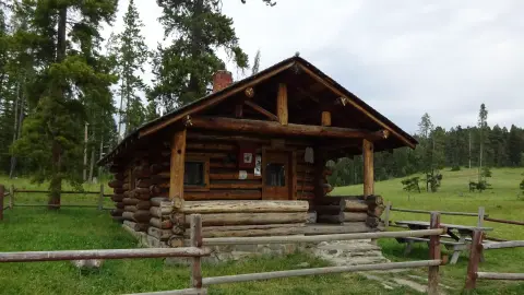 A rustic log cabin stands in a grassy clearing with a wooden fence in the foreground.