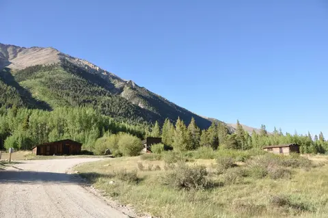 A dirt road leading past wooden cabins toward a large mountain slope.