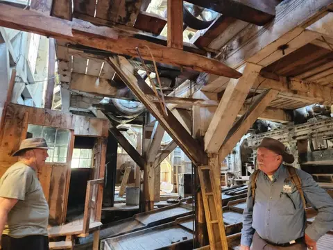 Two men stand inside a rustic wooden structure filled with old machinery and timber framing.