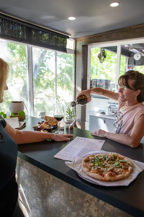 A woman pours wine into a glass at an indoor counter where pizza and snacks are served.