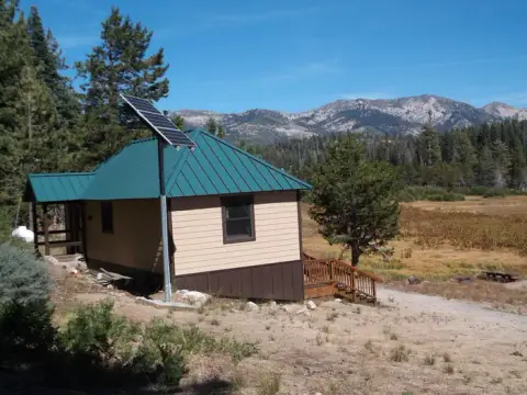 A cabin with a solar panel and green roof sits in a landscape with mountains.