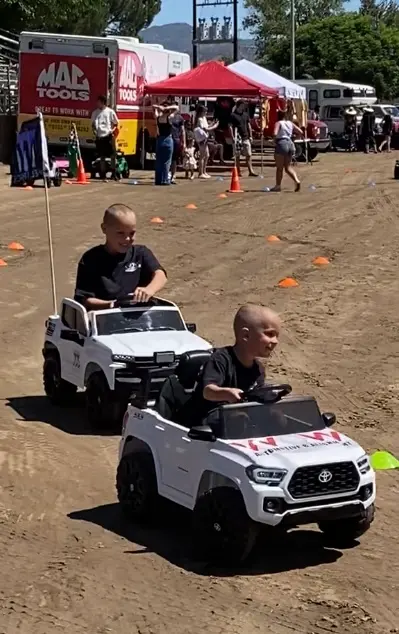 Two children drive toy electric vehicles outdoors on a dirt track.
