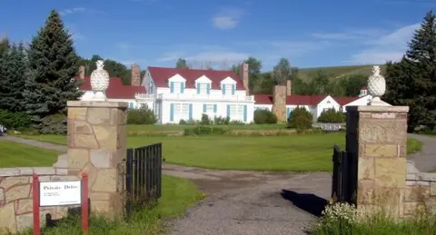 A view of a large white house behind stone pillars and a private drive entrance.