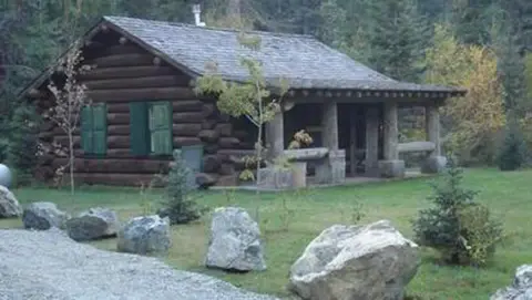 A log cabin with a covered porch sits in a clearing surrounded by trees.