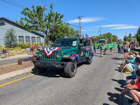 A decorated green Jeep drives down a street during a parade with people watching.
