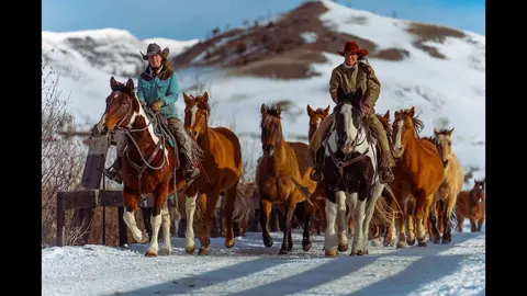 Two riders guide a herd of horses along a snowy path with mountains in the background.