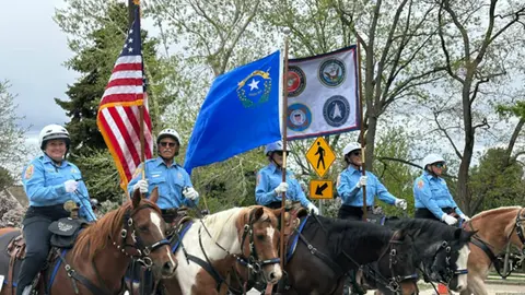 A row of people in uniform ride horses carrying flags outdoors.