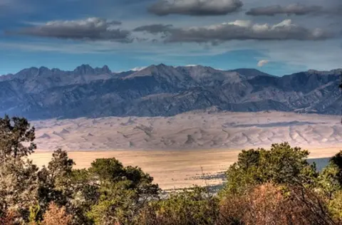 A landscape view showing distant mountains, a wide plain, and trees in the foreground under a cloudy sky.