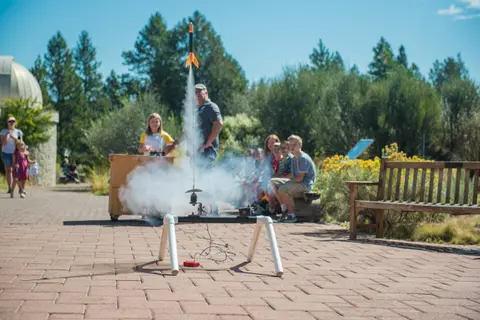 A model rocket launches from a stand on a brick plaza while people watch.