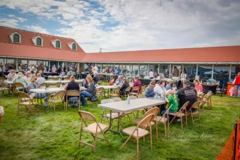 People gather at tables on the lawn outside a long red-roofed building.