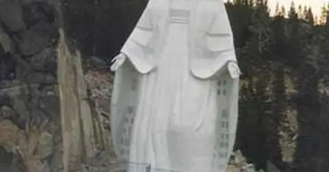 A large white statue stands outdoors near rocky terrain and trees.