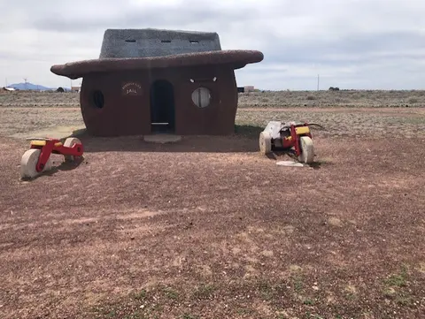 A whimsical building shaped like a hat with stone-like vehicles parked outside in a desert landscape.