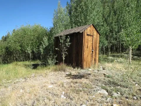 A small wooden shed stands outdoors surrounded by green trees.
