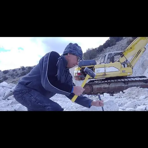 A man kneeling to strike a chisel with a mallet near an excavator.
