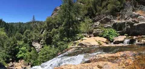 A small waterfall cascades over rocks surrounded by trees and mountains.