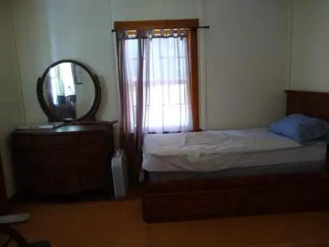 An interior shot of a bedroom with a single bed and a dresser with a round mirror.