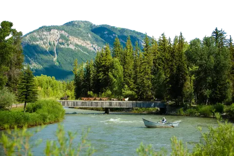 A boat navigates a river beneath a wooden bridge where horses cross, with mountains in the background.