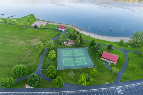 An aerial view of a riverside park with tennis courts, a playground, and walking paths.