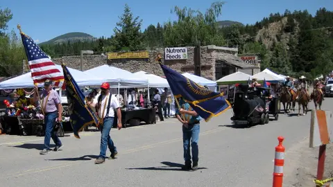 People carry flags in a parade down a street with tents and stores.