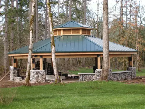 A wooden gazebo with a green metal roof stands in a wooded area.