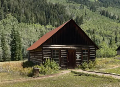 A rustic wooden cabin with a reddish roof sits in a clearing surrounded by a dense forest and mountains.
