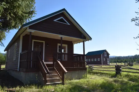 A brown wooden cabin with a porch stands in a grassy field with trees and another building in the background.
