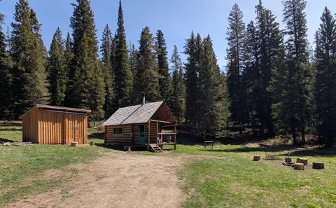 A small log cabin stands in a clearing surrounded by a dense forest of evergreen trees.