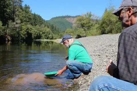 Two men pan for gold in a river with forested mountains in the background.