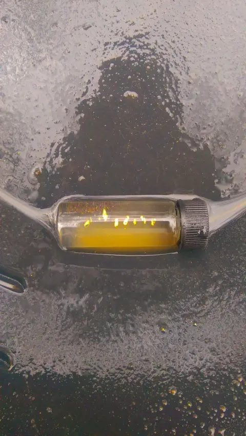 A small vial containing gold flakes and liquid rests on a dark wet surface.