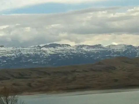 A scenic landscape view of snow-capped mountains behind hills and a body of water under a cloudy sky.