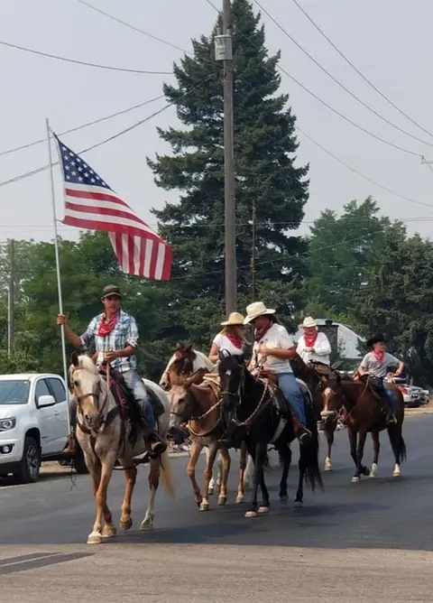 A group of people ride horses down a street in a parade, carrying an American flag.