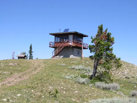 A fire lookout tower on top of a grassy hill under a clear blue sky.