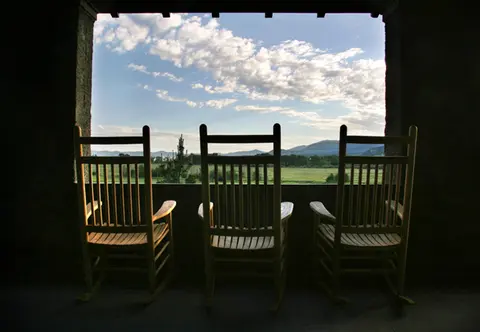 Three wooden rocking chairs sit on a porch looking out at a landscape.