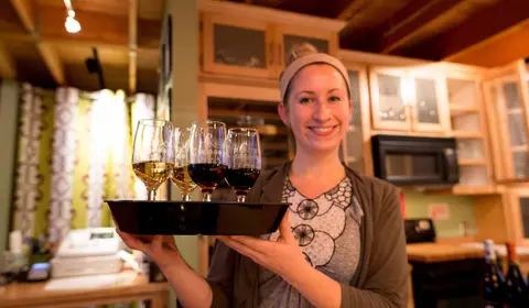 A smiling woman holds a tray with wine glasses inside a building.