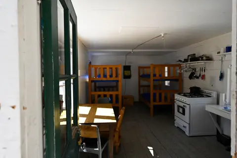 An interior view of a rustic cabin featuring bunk beds, a dining table, and a kitchen area.