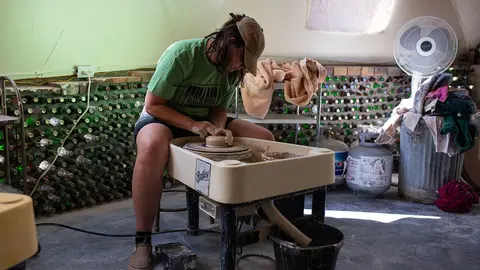 A person works at a pottery wheel inside a room with walls made of glass bottles.