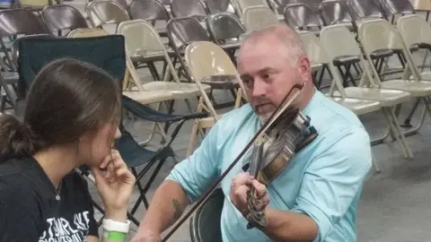 A man plays a violin while a girl watches in a room with many chairs.