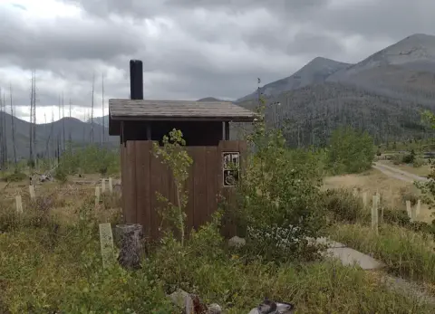 A rustic outhouse stands in a mountainous landscape with cloudy skies.